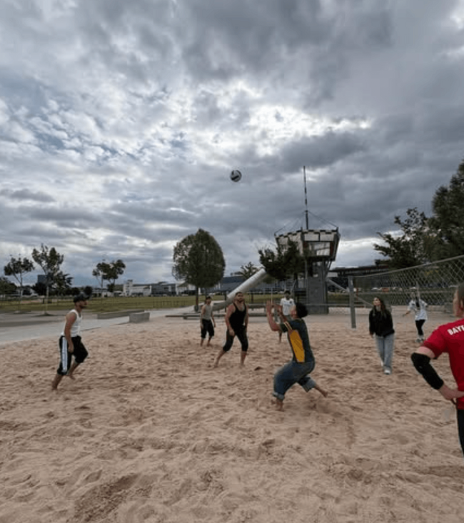 Eine Gruppe von Menschen spielt Volleyball im Sand unter einem bewölkten Himmel während des Projekttags.