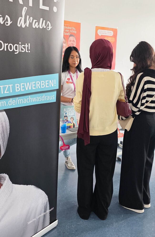 Eine junge Frau erklärt am Informationsstand über die Ausbildung zur Drogistin, während zwei Interessierte zuhören.