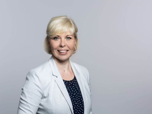 Eine lächelnde Frau mit blonden Haaren trägt einen weißen Blazer vor einem hellgrauen Hintergrund.