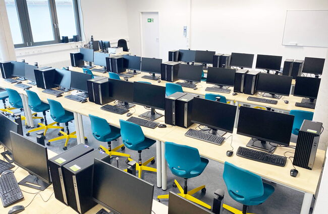 Ein moderner Computerraum mit zahlreichen Arbeitsplätzen, ausgestattet mit Monitoren und ergonomischen blauen Stühlen.