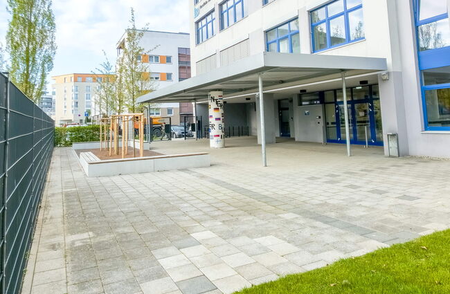 Der Eingang eines modernen Gebäudes mit überdachtem Bereich und angrenzendem, gepflegtem Platz ist zu sehen.