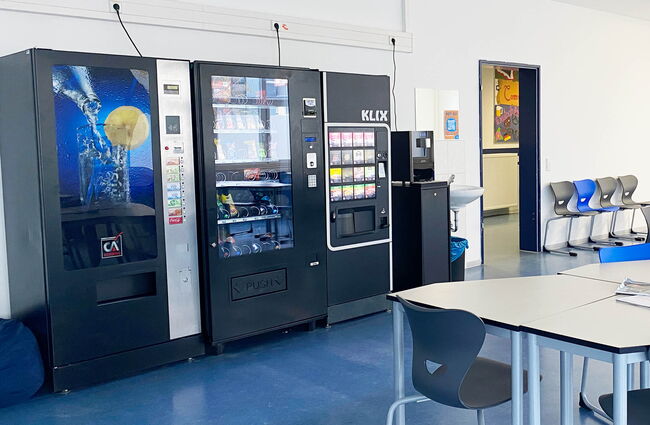 Zwei Snackautomaten stehen in einem hellen Raum, umgeben von modernen Stühlen und einem Waschbecken.