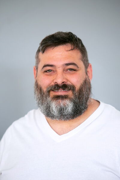 Ein lächelnder Mann mit Vollbart trägt ein weißes T-Shirt vor einem neutralen Hintergrund.