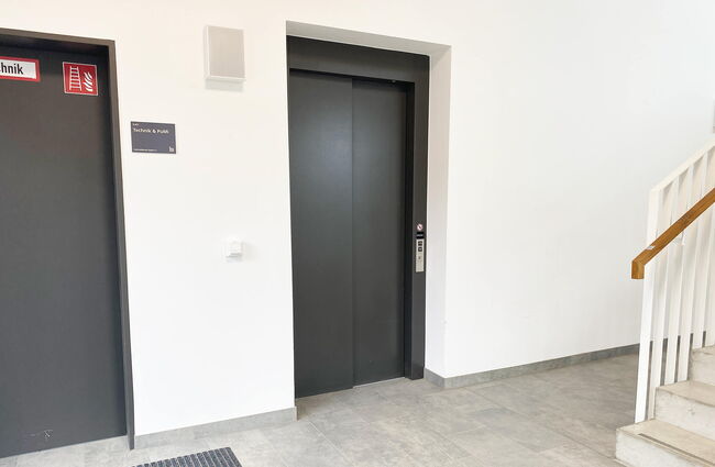 Ein moderner Aufzug steht in einem gepflegten Foyer, umgeben von hellen Wänden und einem Treppenaufgang.