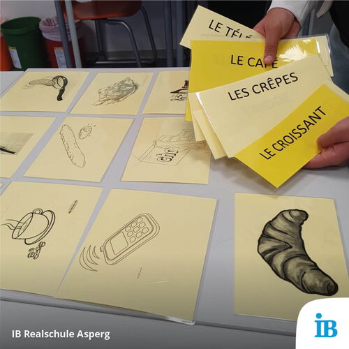 Auf einem Tisch liegen Karten mit französischen Begriffen und Zeichnungen, während jemand eine Karte mit "Le Croissant" hält.