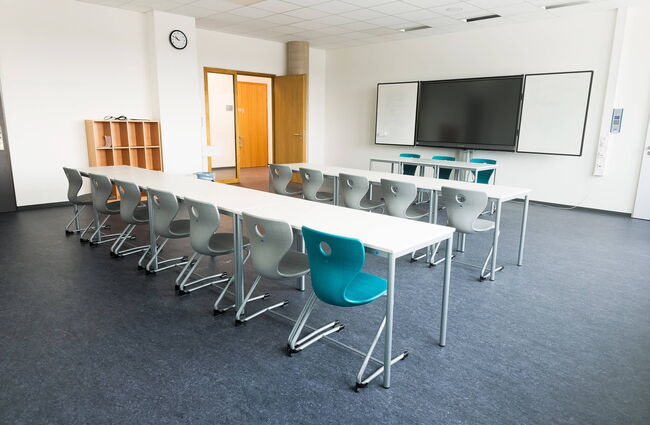 Ein modernes Klassenzimmer mit Tischen, bunten Stühlen und einem großen Bildschirm an der Wand.