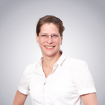 Eine lächelnde Frau in weißem Hemd steht vor einem grauen Hintergrund und strahlt Professionalität aus.