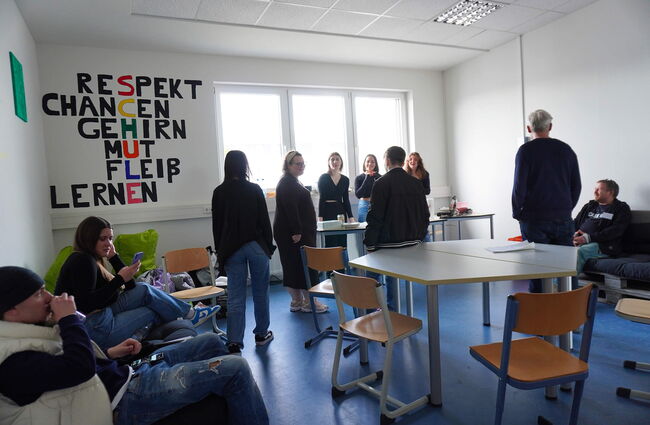 In einem Schulraum stehen Schüler und Lehrer zusammen, während an der Wand inspirierende Begriffe wie „RESPEKT“ und „CHANCEN“ hängen.