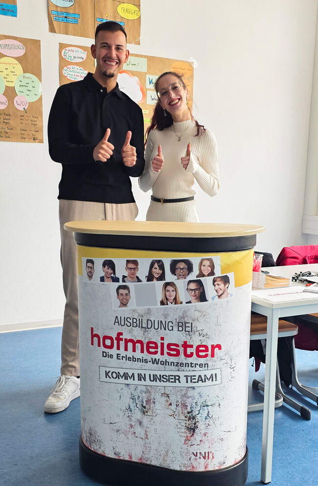 Zwei fröhliche junge Erwachsene stehen am Tisch für die Ausbildung bei Hofmeister und zeigen Daumen nach oben.