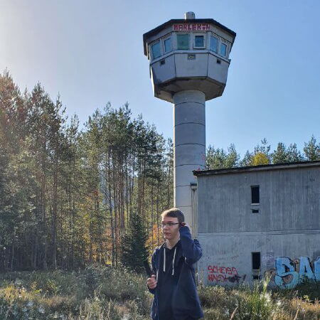 Ein Mann steht vor einem alten Wachturm und einem verlassenen Gebäude in Ludwigswinkel, umgeben von Bäumen und Graffiti.