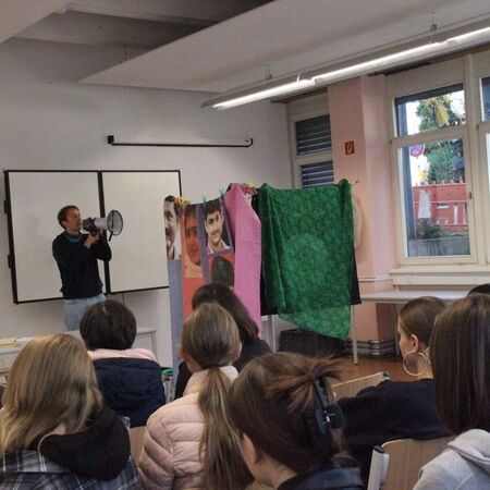 Ein Mann spricht durch ein Megafon vor einer Klasse, während bunte Tücher und Poster im Hintergrund hängen.