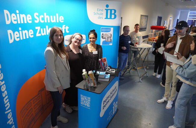 Drei freundliche Frauen stehen an einem Informationsstand, während interessierte Besucher mit Unterlagen in der Schlange warten.