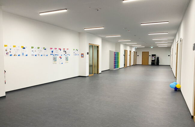 Ein heller Flur im Neubau mit bunten Plakaten an der Wand und einem Getränkeautomaten am Ende des Raumes.