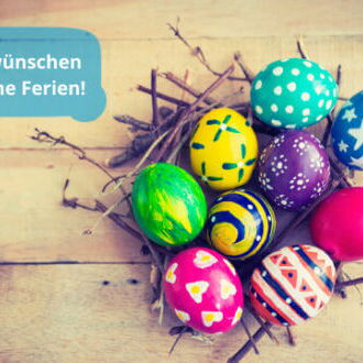Bunte Ostereier liegen auf einem Holztisch neben einem Schild mit der Aufschrift "Wir wünschen schöne Ferien".