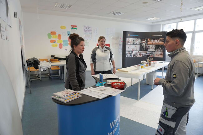 Drei Personen diskutieren in einem Klassenraum an einem blauen Infostand über Ausbildungsprogramme.