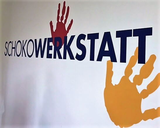 Ein Schild mit der Aufschrift "Schokowerkstatt" und bunten Handabdrücken ziert die Wand.