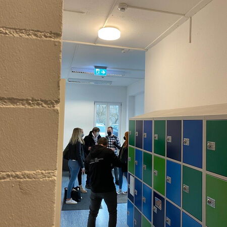 Eine Gruppe von Menschen unterhält sich neben bunten Schließfächern in einem Flur mit Notausgangsschild.