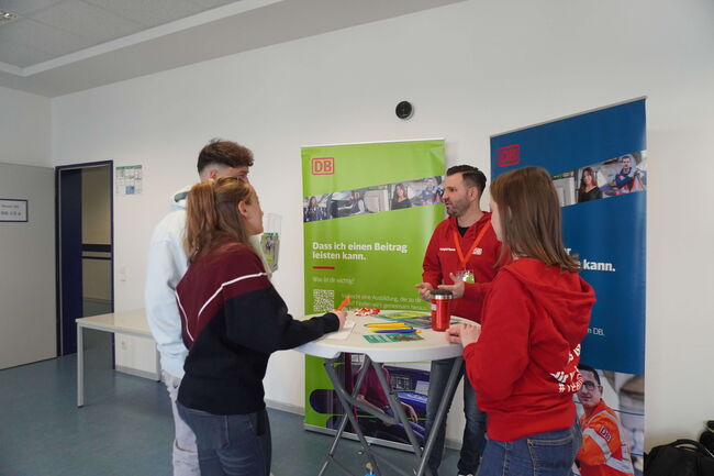 Zwei junge Menschen informieren sich an einem Stand mit DB-Bannern bei zwei Mitarbeitern in roter Kleidung.