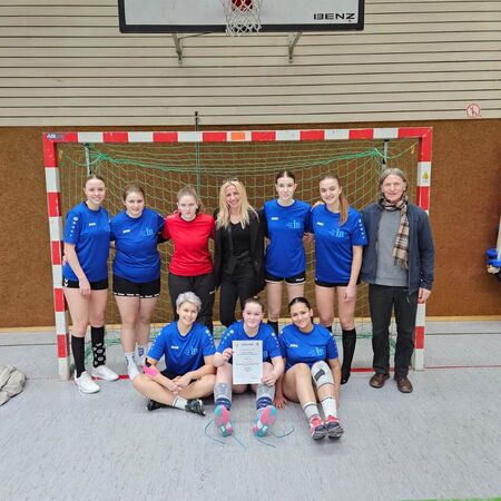 Eine Gruppe von Handballspielerinnen posiert in blauen Trikots vor einem Tor in einer Sporthalle mit einer Urkunde.