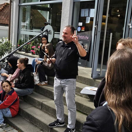 Eine Gruppe junger Menschen sitzt auf Treppenstufen vor einem Café, während ein Mann gestikulierend spricht.