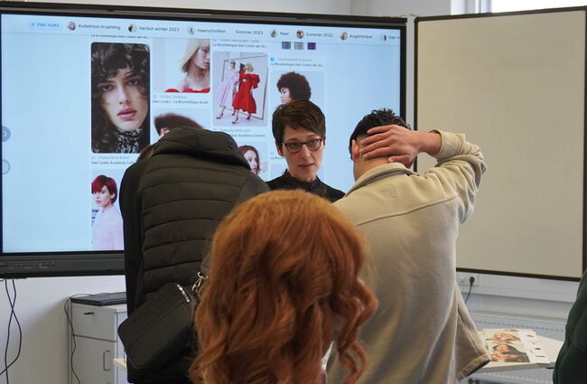 Eine Lehrkraft erklärt den Teilnehmern verschiedene Haarlooks anhand einer digitalen Präsentation in einem modernen Klassenraum.