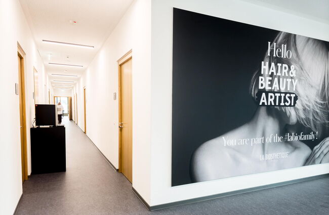 Ein heller Flur mit einer großen Wandgrafik, die „Hello Hair & Beauty Artist“ zeigt, und einem Computer auf einem Tisch.