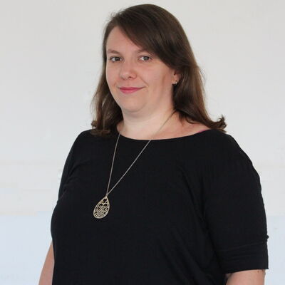 Eine Frau in schwarzem Oberteil lächelt leicht, während sie einen filigranen Anhänger trägt, vor einem neutralen Hintergrund.