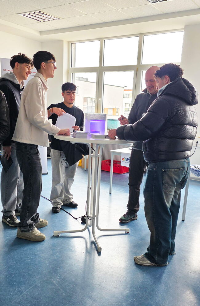 Eine Gruppe junger Leute steht an einem Tisch und diskutiert über ein leuchtendes Gerät bei einer Informationsveranstaltung.