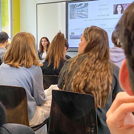 Eine Frau hält in einem Klassenzimmer vor Schülern einen Vortrag über den Bundestag, während eine Präsentation läuft.