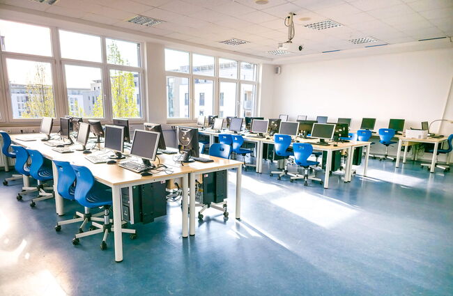 Ein moderner Computerraum mit mehreren Arbeitsplätzen, blauen Stühlen und großen Fenstern für viel Tageslicht.