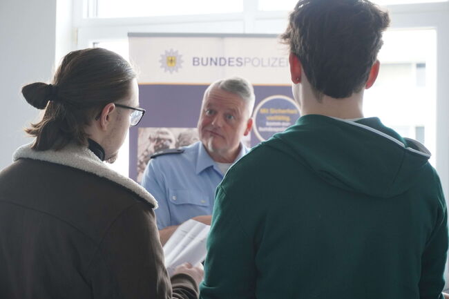 Ein Polizist der Bundespolizei spricht mit zwei jungen Männern vor einem Informationsstand.