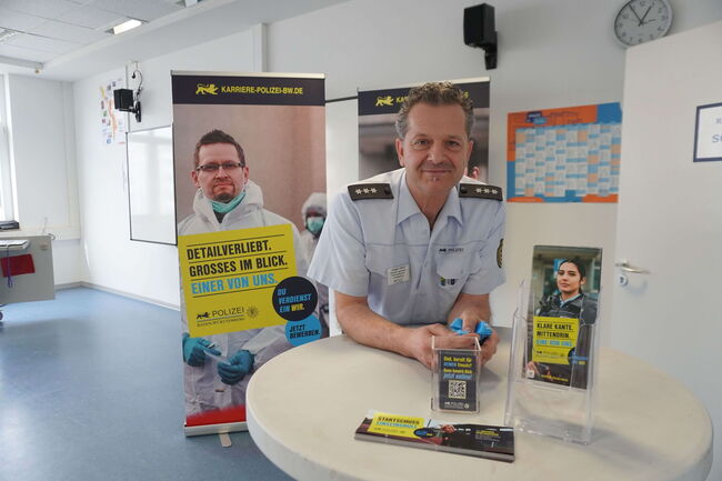 Ein Polizist steht an einem Tisch mit Informationsmaterialien und einem großen Werbebanner zur Polizeikarriere.