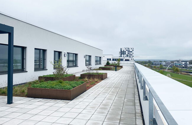 Die Dachterrasse eines Neubaus zeigt bepflanzte Beete und einen weiten Blick auf die umliegende Landschaft.