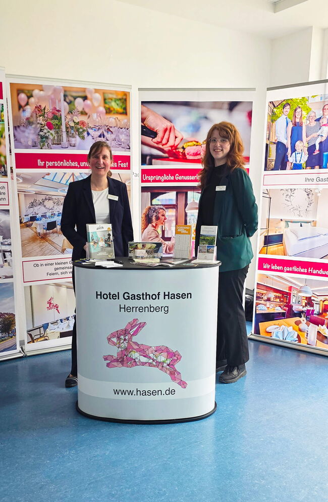 Zwei freundliche Mitarbeiterinnen stehen am Stand des Hotel Gasthof Hasen und präsentieren Informationsmaterial.