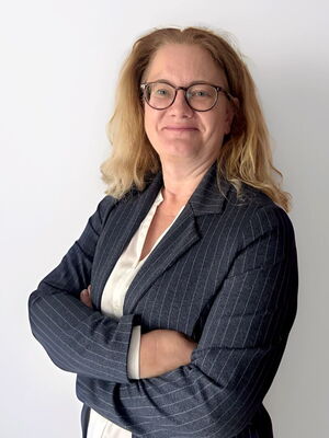 Eine Frau mit Brille und blonden Haaren trägt einen dunkel gestreiften Blazer und lächelt mit verschränkten Armen.