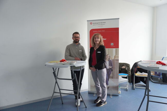 Zwei Personen stehen an einem Infostand der Bundesagentur für Arbeit in einem hellen Raum.