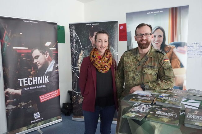 Ein Soldat und eine Frau stehen lächelnd vor einem Bundeswehr-Infostand mit Werbebannern und Broschüren.