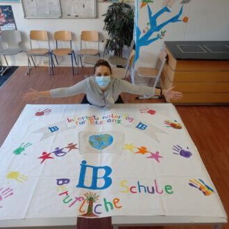 Eine Person mit Maske präsentiert ein buntes Poster mit Handabdrücken und der Aufschrift "IB Schule" in einem hellen Raum.