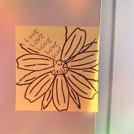 Ein gelber Zettel mit einer gezeichneten Blume und dem Wort "Love" mehrfach darauf steht an einer Wand.