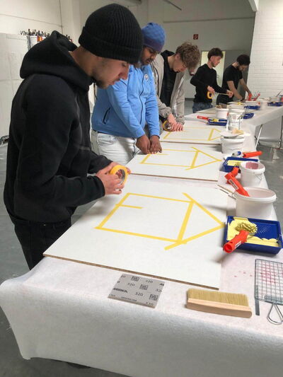 Fünf Personen arbeiten konzentriert an Tafel mit gelbem Klebeband, um geometrische Muster für ein kreatives Projekt zu erstellen.