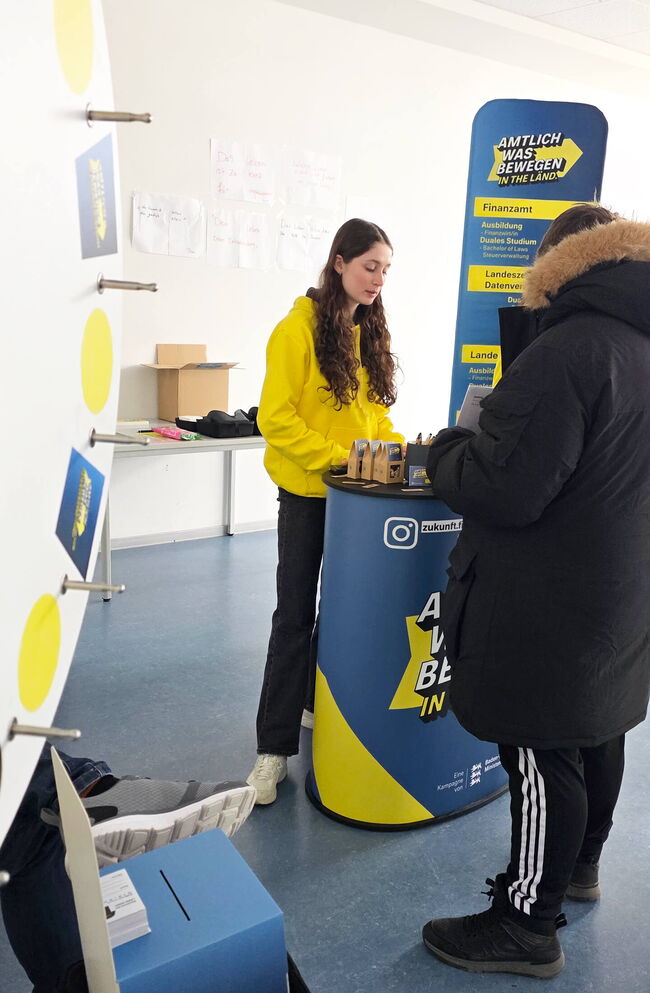 Eine junge Frau in einem gelben Pullover berät einen Besucher am Informationsstand über Ausbildungsangebote im Finanzamt.
