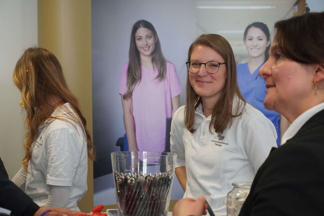 Frauen in einem medizinischen Umfeld lächeln bei einer Informationsveranstaltung neben einem Stand mit Stiften.