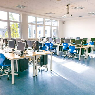 Ein moderner Computerraum mit mehreren Arbeitsplätzen, blauen Stühlen und großen Fenstern für viel Tageslicht.