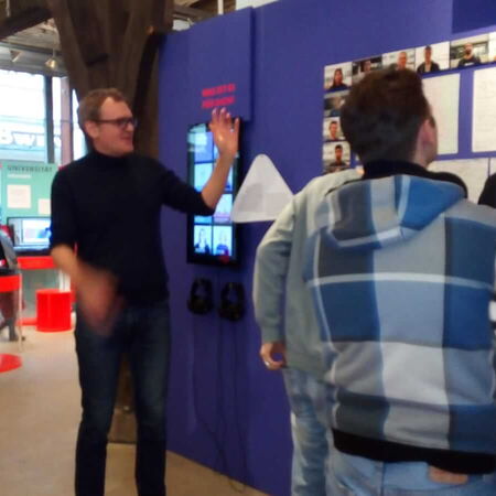 Besucher betrachten eine Ausstellung in einem modernen Raum, während ein Mann gestikulierend etwas erklärt.