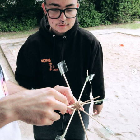 Eine Person im schwarzen Hoodie hält ein selbstgebautes Spielgerät aus Korken und Stäben auf einem Sandspielplatz.