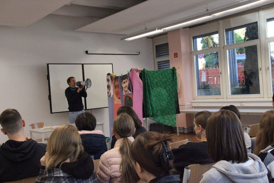Ein Mann spricht durch ein Megafon vor einer Klasse, während bunte Tücher und Poster im Hintergrund hängen.