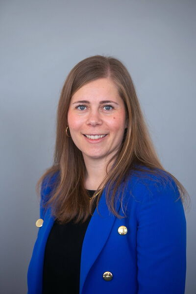 Eine lächelnde Frau in einem blauen Blazer posiert vor einem grauen Hintergrund für ein Portraitfoto.