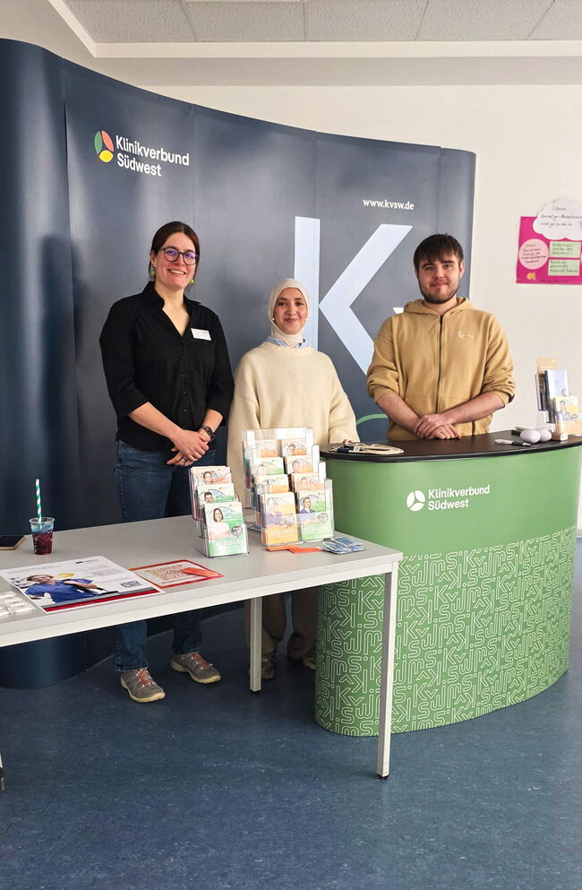 Drei Mitarbeiter des Klinikverbund Südwest stehen an einem Informationsstand mit Flyern und Materialien.