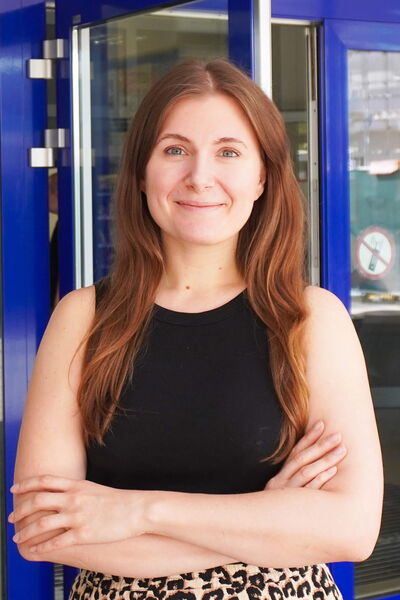 Eine lächelnde Frau mit langen, braunen Haaren steht vor einer blauen Tür und hat die Arme verschränkt.