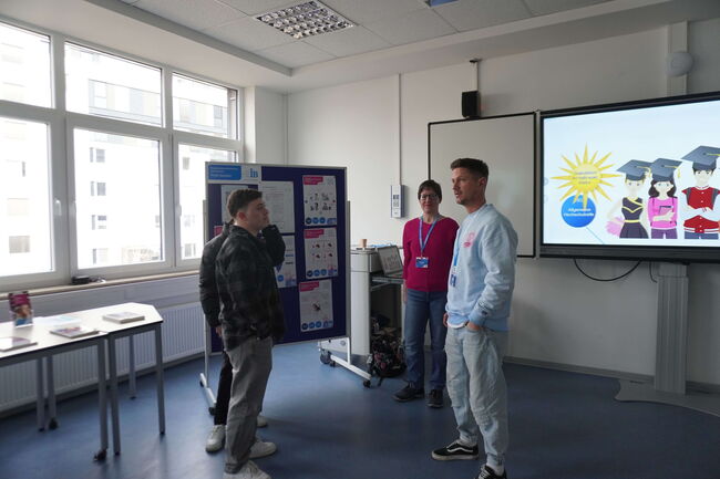 Drei Personen diskutieren vor einem Bildschirm in einem Klassenzimmer über Bildungswege und Präsentationen.